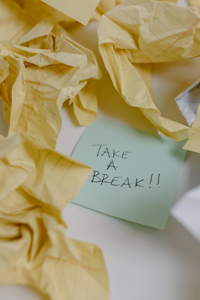 Une note autocollante verte avec les mots "take a break" à côté de plusieurs feuilles de papier jaune froissées.