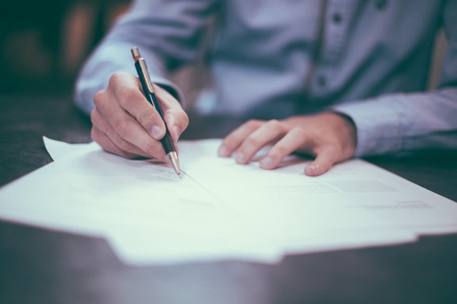 A person with a blue full-sleeve shirt writes on a sheet of paper with a black pen.