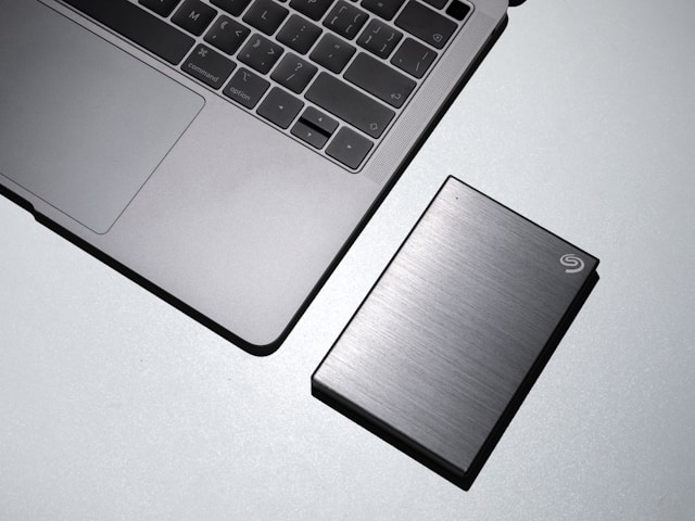 MacBook abu-abu di samping solid-state drive eksternal Seagate berwarna abu-abu.
