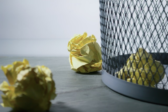 Een close-up van een grijze vuilnisbak met verfrommeld geel papier erin en op de vloer.