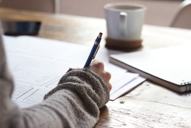 Una persona con un maglione marrone usa una penna blu per disegnare su un foglio di carta.
