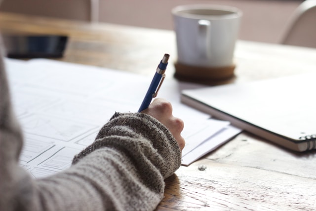 A person with a brown sweater uses a blue pen to draw on a sheet of paper.
