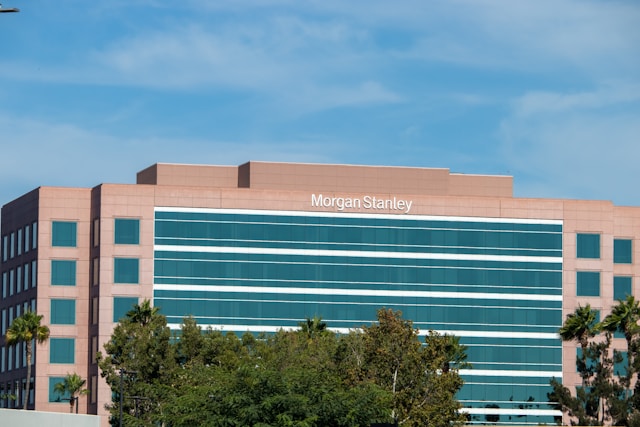 Ein hellbraunes Bürogebäude mit einem weißen "Morgan Stanley"-Schild hinter mehreren Bäumen.