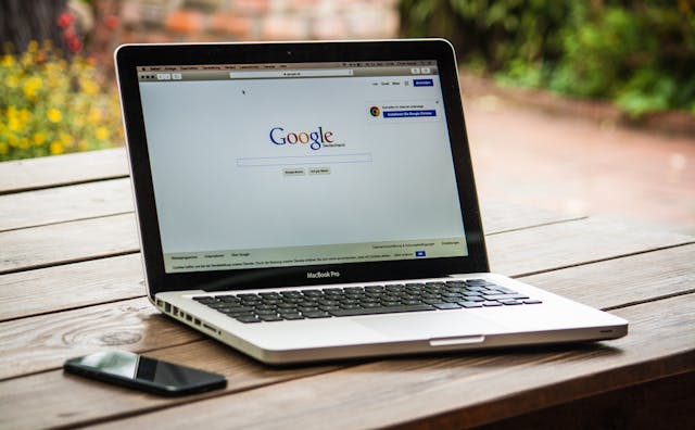 Google-Suche auf einem grauen MacBook Pro und einem schwarzen Smartphone auf einem Holztisch.