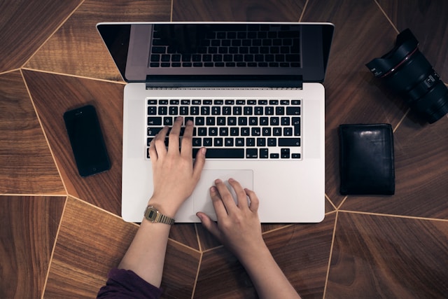 O persoană folosește tastatura și trackpad-ul MacBook Pro pe o masă maro cu portofelul și obiectivul aparatului foto.