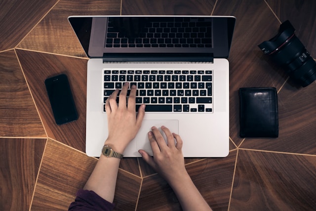 한 사람이 지갑과 카메라 렌즈를 들고 갈색 테이블 위에서 MacBook Pro의 키보드와 트랙패드를 사용하고 있습니다.