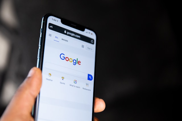 Eine Person hält ein schwarzes iPhone, auf dessen Display die Startseite der Google-Suche angezeigt wird.