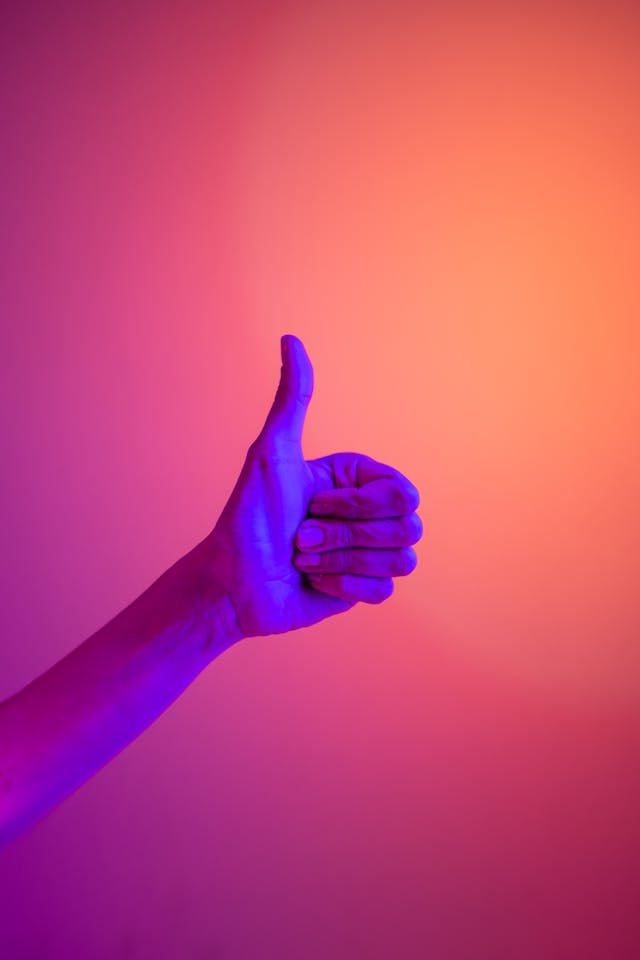 A person shows a thumbs-up in front of an orange wall.