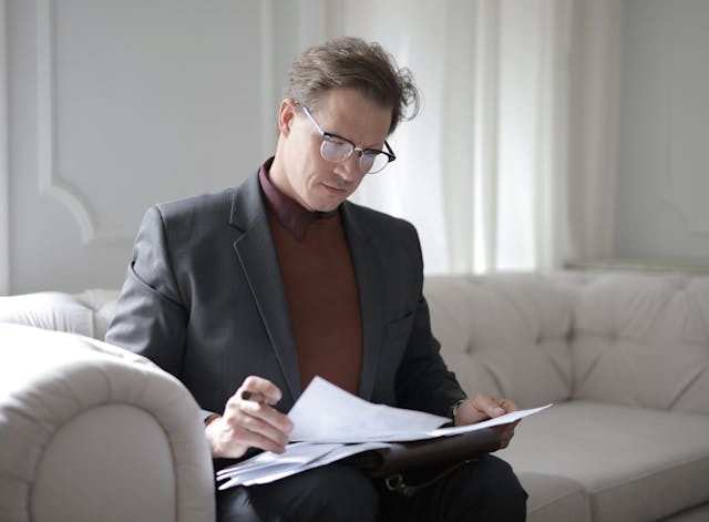 Un homme en costume gris et chemise marron est assis sur un canapé blanc et examine des feuilles de papier.