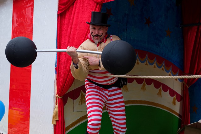 A man with striped overalls and a light brown jacket lifts a barbell with one hand.