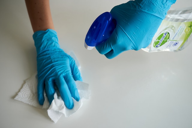 Een persoon met blauwe handschoenen houdt een spuitfles vast en veegt een oppervlak af met een tissue.