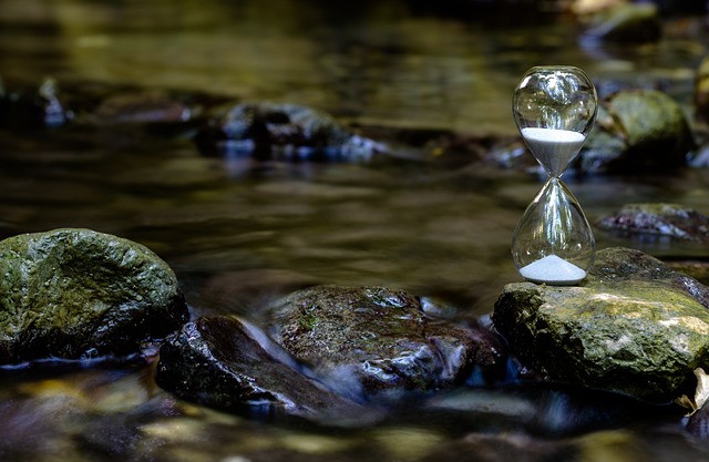 An hourglass with white sand on a rock above a water body.