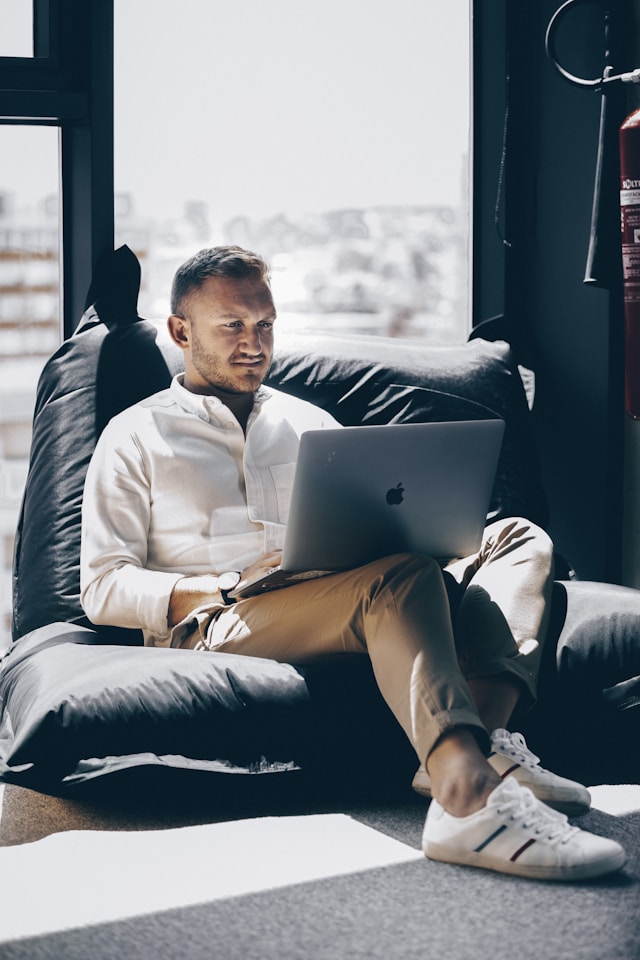 흰색 셔츠와 갈색 바지를 입은 남성이 검은색 소파에 앉아 회색 MacBook Pro를 사용하고 있습니다.