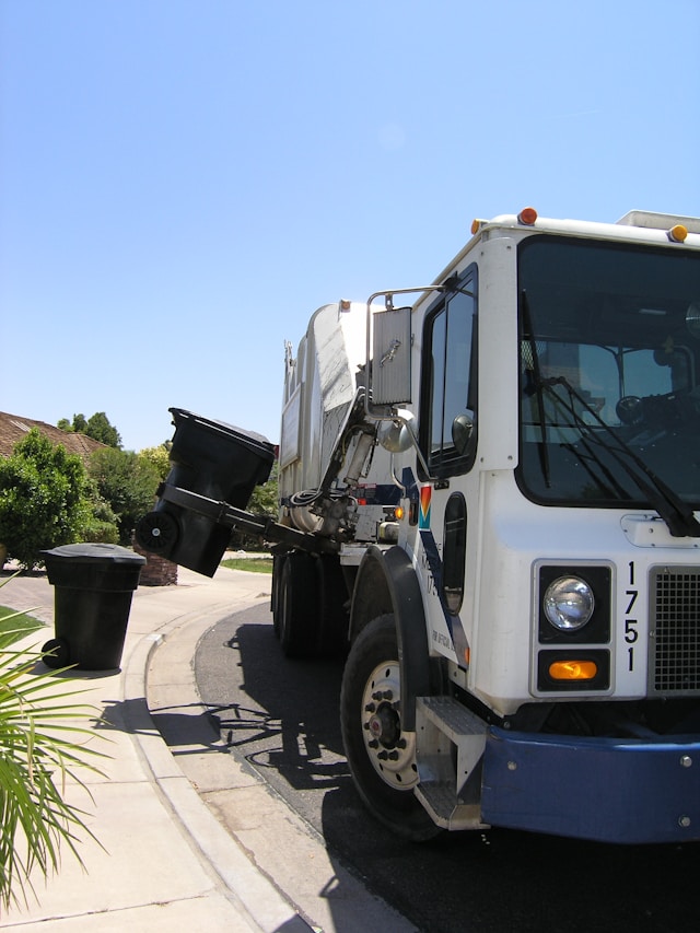 Un camión de la basura blanco recoge un cubo de basura negro.