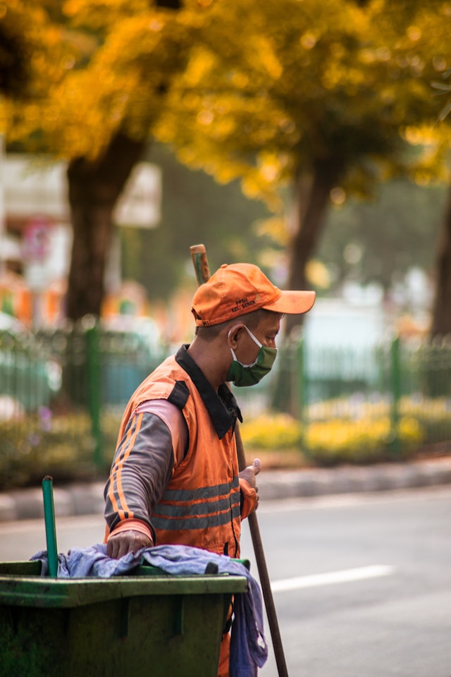 オレンジ色の帽子と視認性の高いベストを着たゴミ収集作業員が、棒と緑色のゴミ箱を持っている。