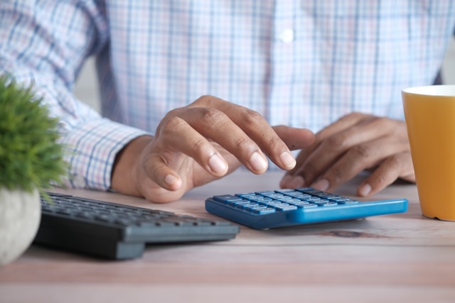 Una persona con una camisa a rayas azules y rojas utiliza una calculadora azul sobre una mesa marrón.