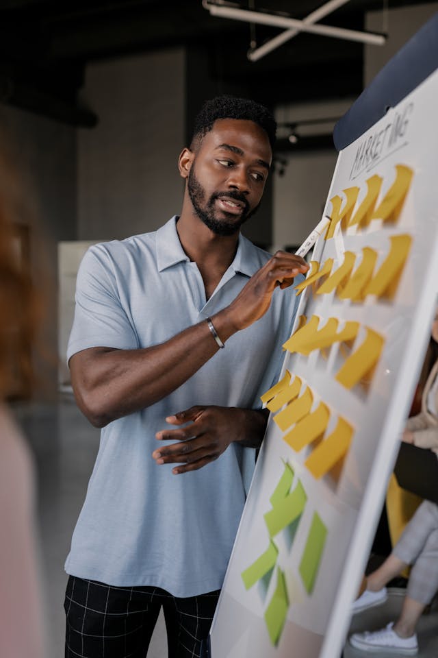 Un homme à la chemise grise montre un tableau blanc sur lequel sont collées de nombreuses notes.