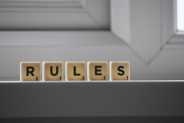 Gros plan sur le mot "rules" dans cinq tuiles de Scrabble sur un mur blanc.