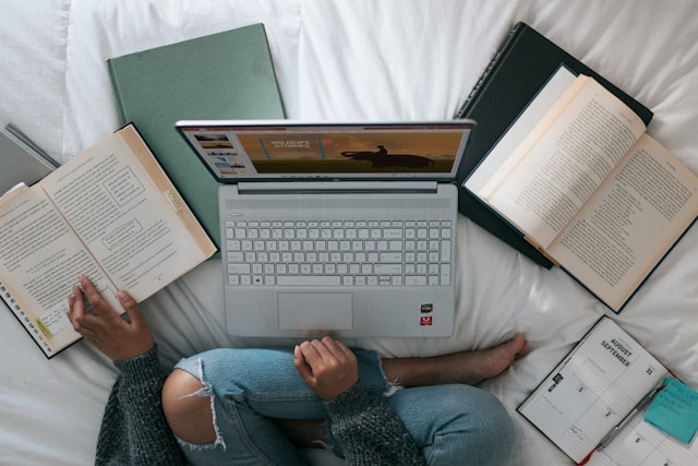 شخص يجلس على سرير أبيض، ويضع يده على كتاب، ويستخدم جهاز كمبيوتر محمول رمادي اللون