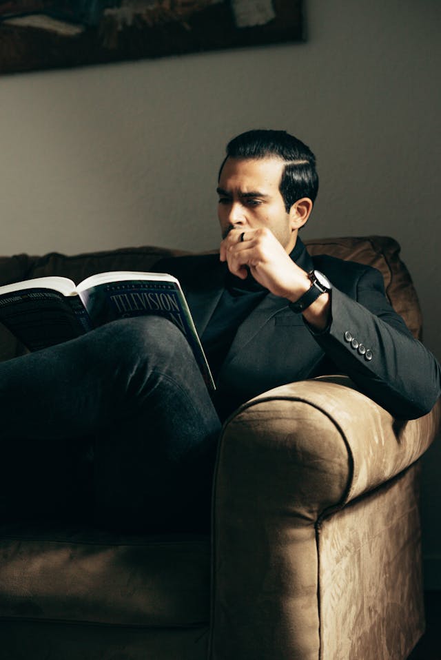 黒いスーツを着た人物が茶色のソファに座って本を読んでいる。
