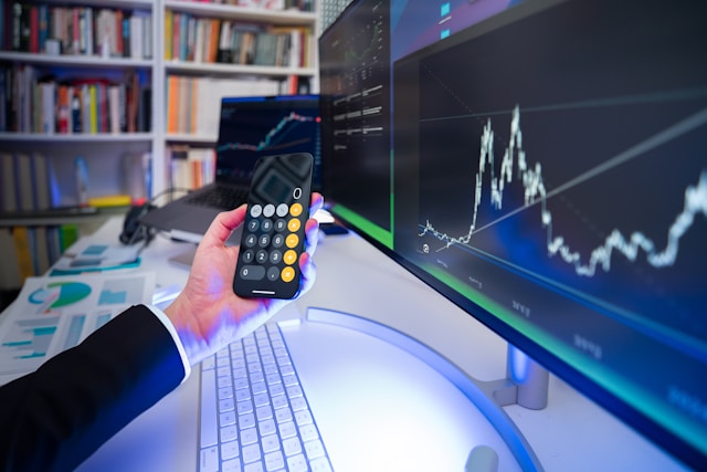 A person holds a black iPhone with the calculator app open next to a monitor with a graph.