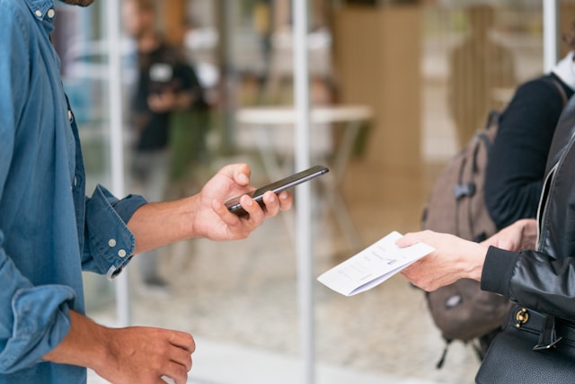 Eine Person hält ein schwarzes Smartphone in der Hand und richtet es auf eine Person mit einem Blatt Papier.