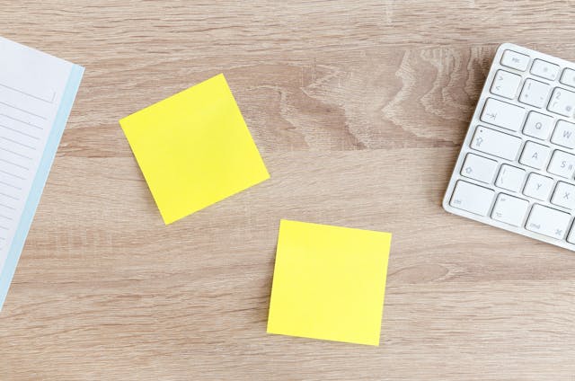 Two yellow sticky notes between a notebook and a white keyboard on a brown surface.