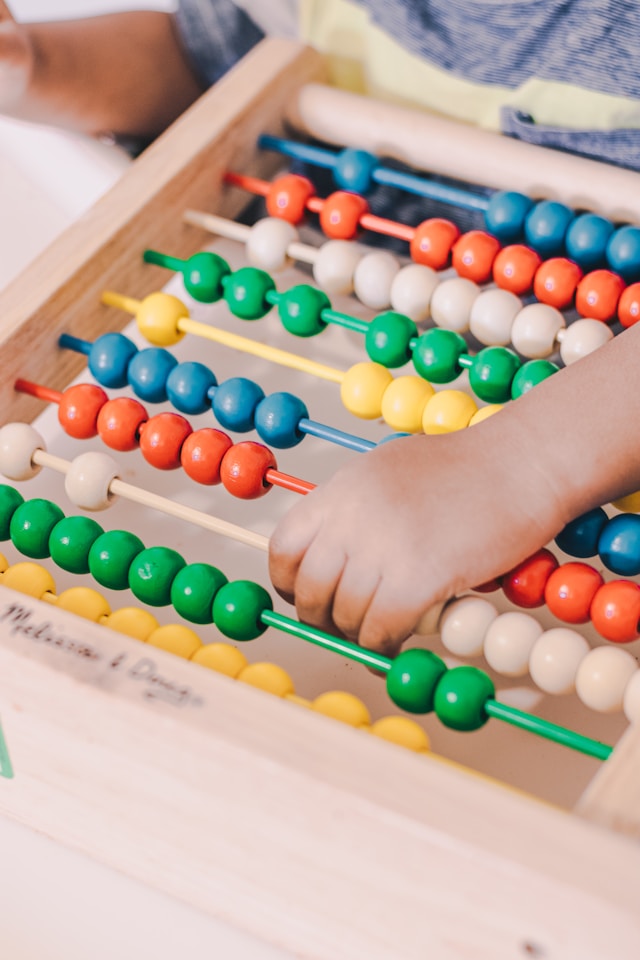 Een hand houdt een houten staaf vast van een bruin telraam met gele, groene, blauwe, rode en witte kralen.