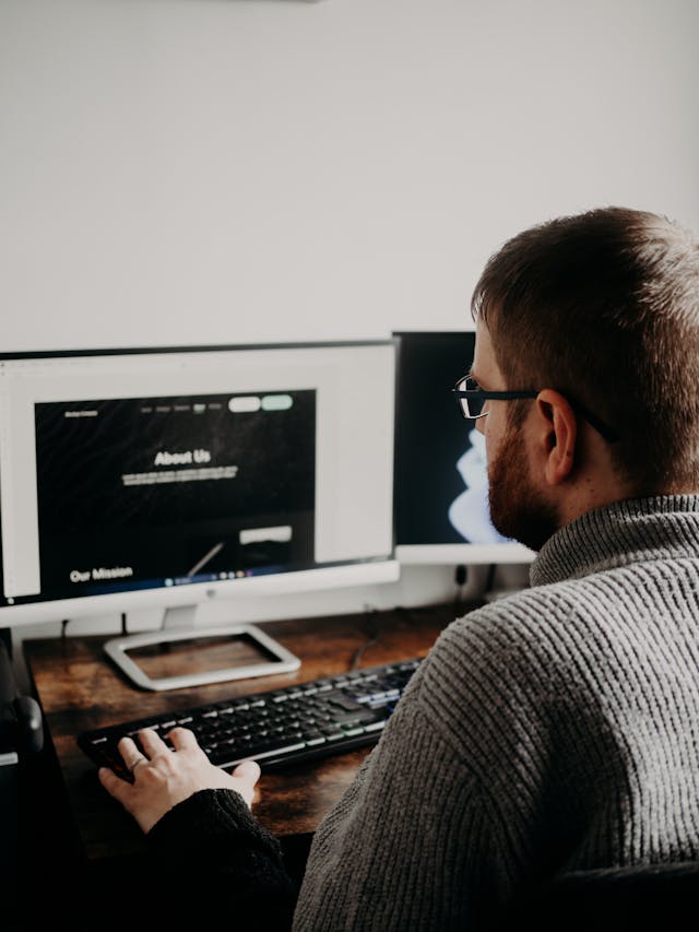 Um homem com uma camisola cinzenta olha para um ecrã e utiliza um teclado preto.