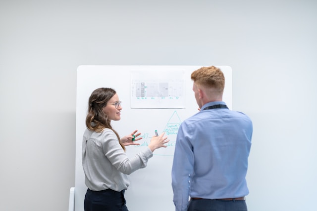 Een vrouw met een lichtgrijs overhemd en een man met een blauw overhemd converseren voor een whiteboard. 