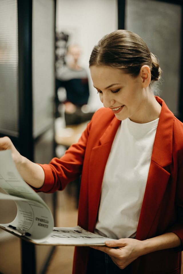 Une femme en tailleur rouge et chemise blanche tient un presse-papiers et consulte un document.