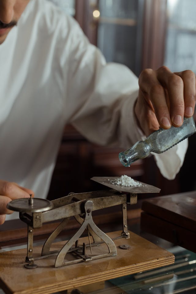 Une personne portant une chemise blanche pèse une poudre blanche contre un petit poids sur une vieille balance.