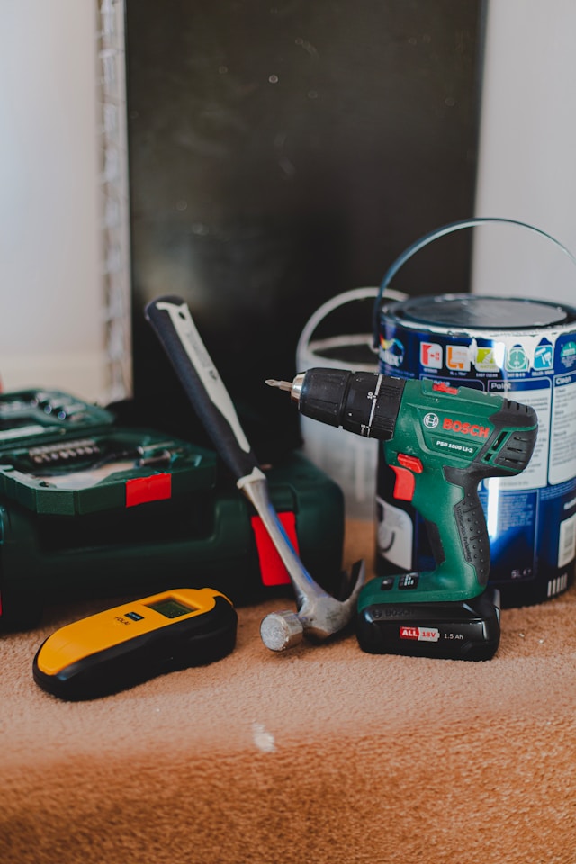 A collection of a battery-powered drill, a hammer, a paint can, and other tools on an orange carpet.