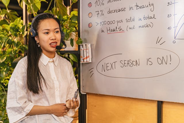 A woman with a headset and a white shirt stands next to a whiteboard.