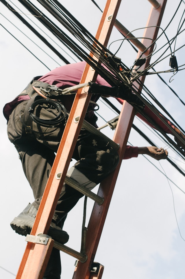 An electrician with protective gear on a ladder fixes various electrical wires.