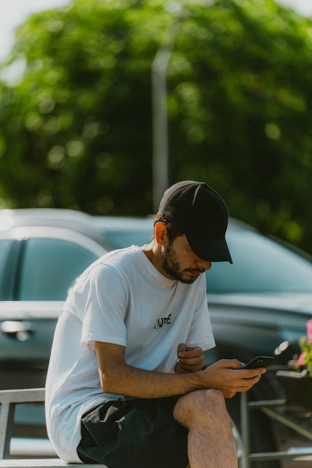 Un bărbat cu un tricou alb, pantaloni scurți negri și o șapcă neagră stă și folosește un smartphone negru.