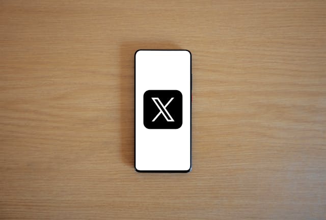 A mobile phone on a wooden surface displays the X logo.