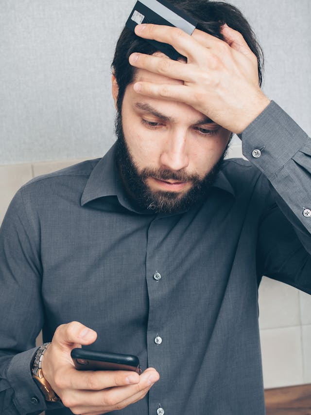 A man with a gray shirt holds a card and looks concerned after he looks at his phone.