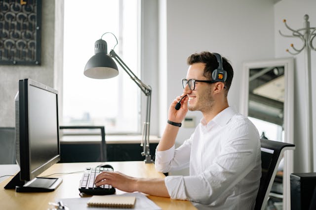 A man with a white shirt holds the microphone on his headset and uses a keyboard.