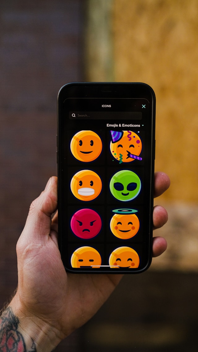 A person holds a black smartphone with multiple emojis on its display.