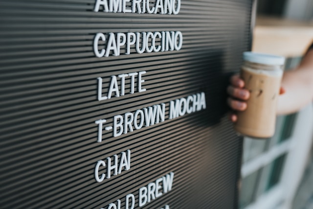 A person holds a cup with a brown liquid next to the names of various beverages on a blackboard.