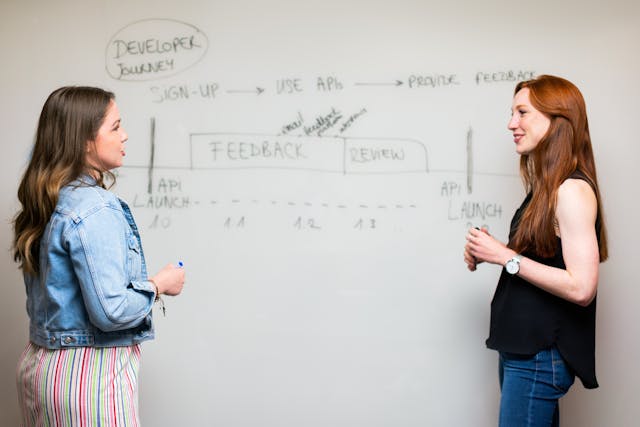 Two women stand next to a whiteboard and talk about APIs.