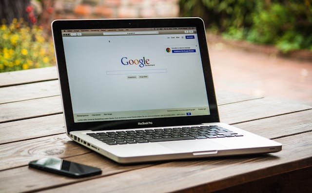 A gray MacBook Pro on a brown wooden table with Google Search on its display next to a black smartphone.