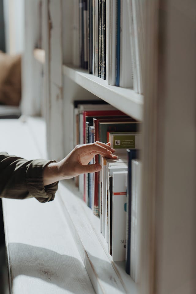 A person with a green sleeve touches a book on a white bookshelf.