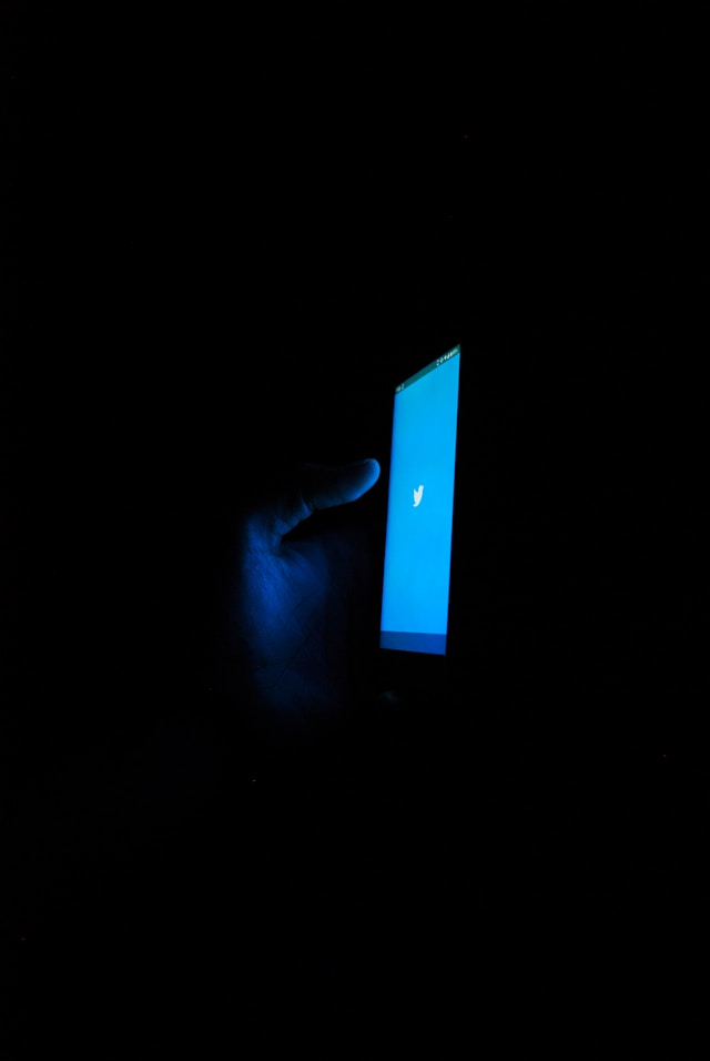 A blue Twitter logo glows on a smartphone screen held by a hand in the dark.