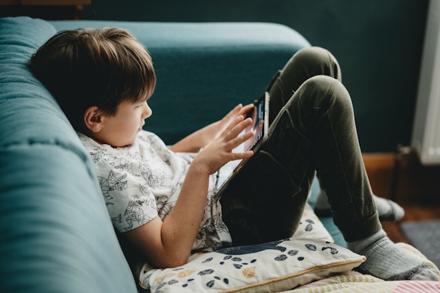 A kid sits on a white cushion on a blue sofa and uses a tablet.