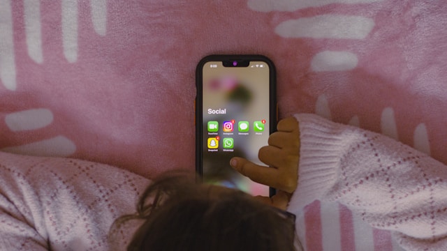 A person lies on a bed with pink sheets and opens the social media folder on a black iPhone.