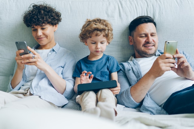 A woman, a child, and a man sit on a bed and use various electronic devices.