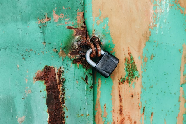 O ușă metalică cu vopseaua verde care se desprinde, ținută împreună de un lacăt negru.