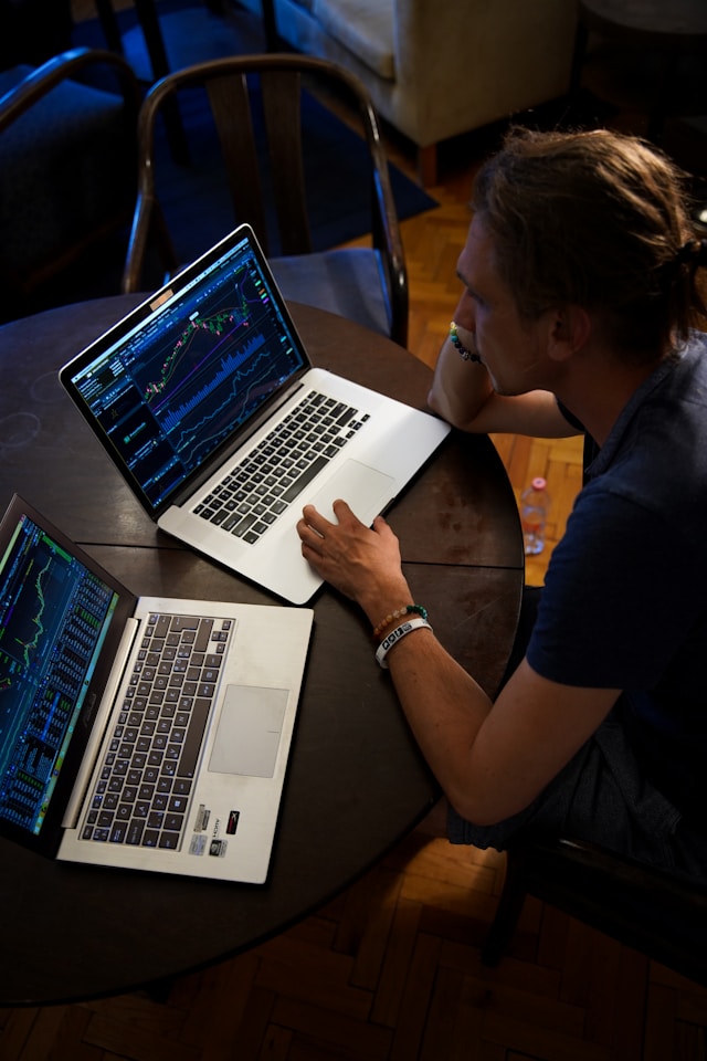 Un bărbat se uită la un grafic pe un Macbook gri, alături de un laptop gri cu Windows. 
